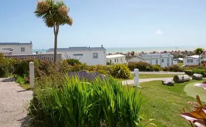 A sunny holiday park with modern mobile homes, lush green lawns, and a distant ocean view.