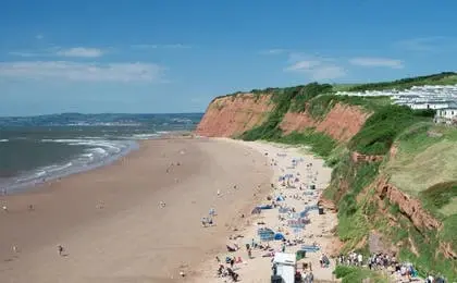 A wide, sandy beach stretching beneath dramatic red cliffs under a bright blue sky.