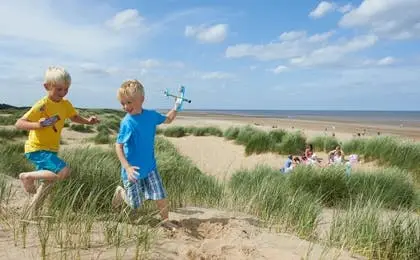 Two young boys running through grassy sand dunes near a beach, with one holding a toy airplane.