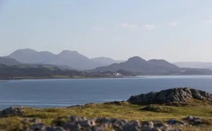 A scenic view of calm blue water with distant mountains under a clear sky, viewed from a grassy, rocky shore.