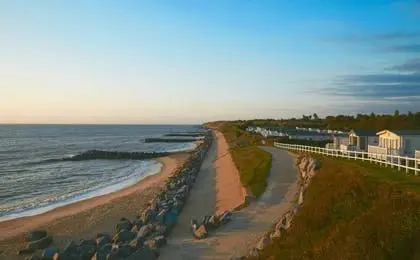 A scenic coastal view featuring a sandy beach, rock breakwaters, a paved path, and holiday homes along the shore.