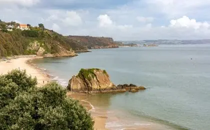 A scenic view of a sandy beach, green cliffs, and a rocky outcrop in the calm sea under a cloudy sky.