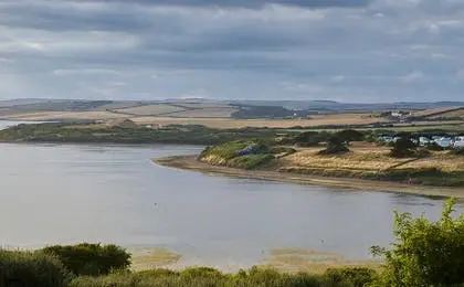 A wide estuary with calm water under a cloudy sky, bordered by green hills and distant fields.