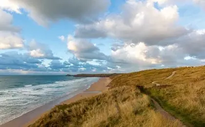 A scenic coastal view with rolling grassy hills meeting a sandy beach and blue ocean under a cloudy sky.