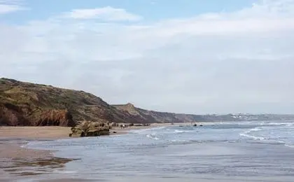 A scenic coastal beach with cliffs, gentle waves, and a cloudy sky.