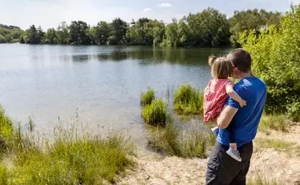 A father holding his young daughter by a scenic lake on a sunny day.