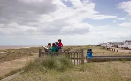 A group of children looking at a display panel near a sandy beach under a cloudy sky.