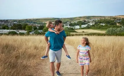 A father giving his son a piggyback ride on a grassy path while a young girl walks alongside them.