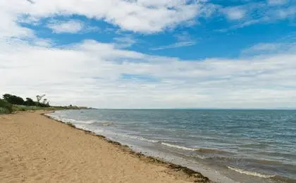 A wide sandy beach meeting calm ocean waters under a partly cloudy blue sky.
