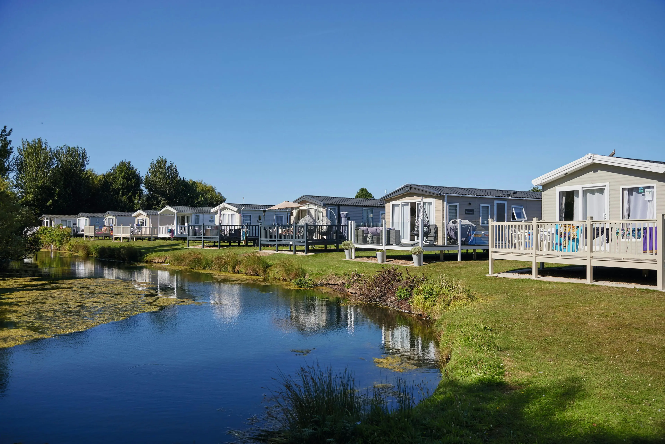 Row of modern static caravans overlooking a serene water canal on a sunny day.