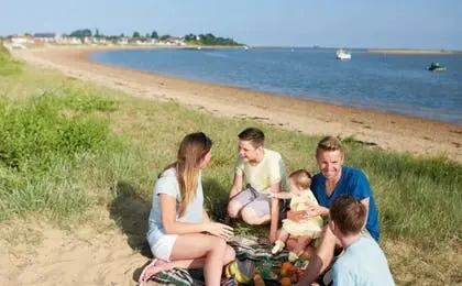 A family enjoying a sunny picnic on a grassy hill overlooking a sandy beach and water.