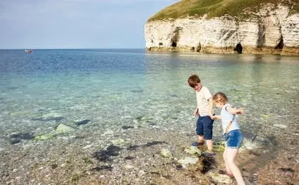 Two young children playing in the shallow clear water near a white chalk cliff.