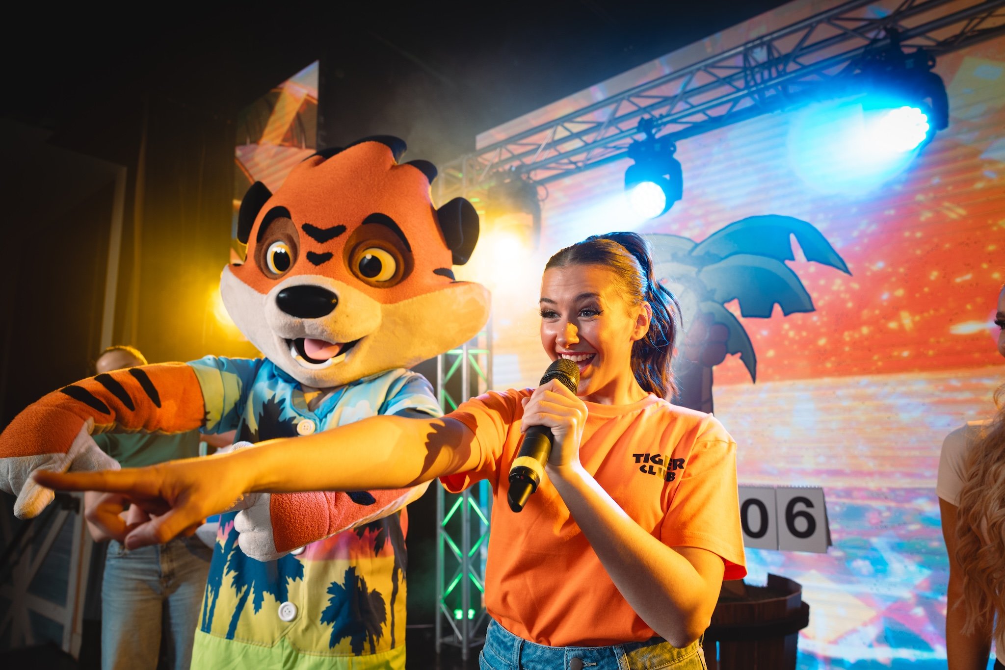 A smiling woman holding a microphone points forward next to a tiger mascot on a brightly lit stage.