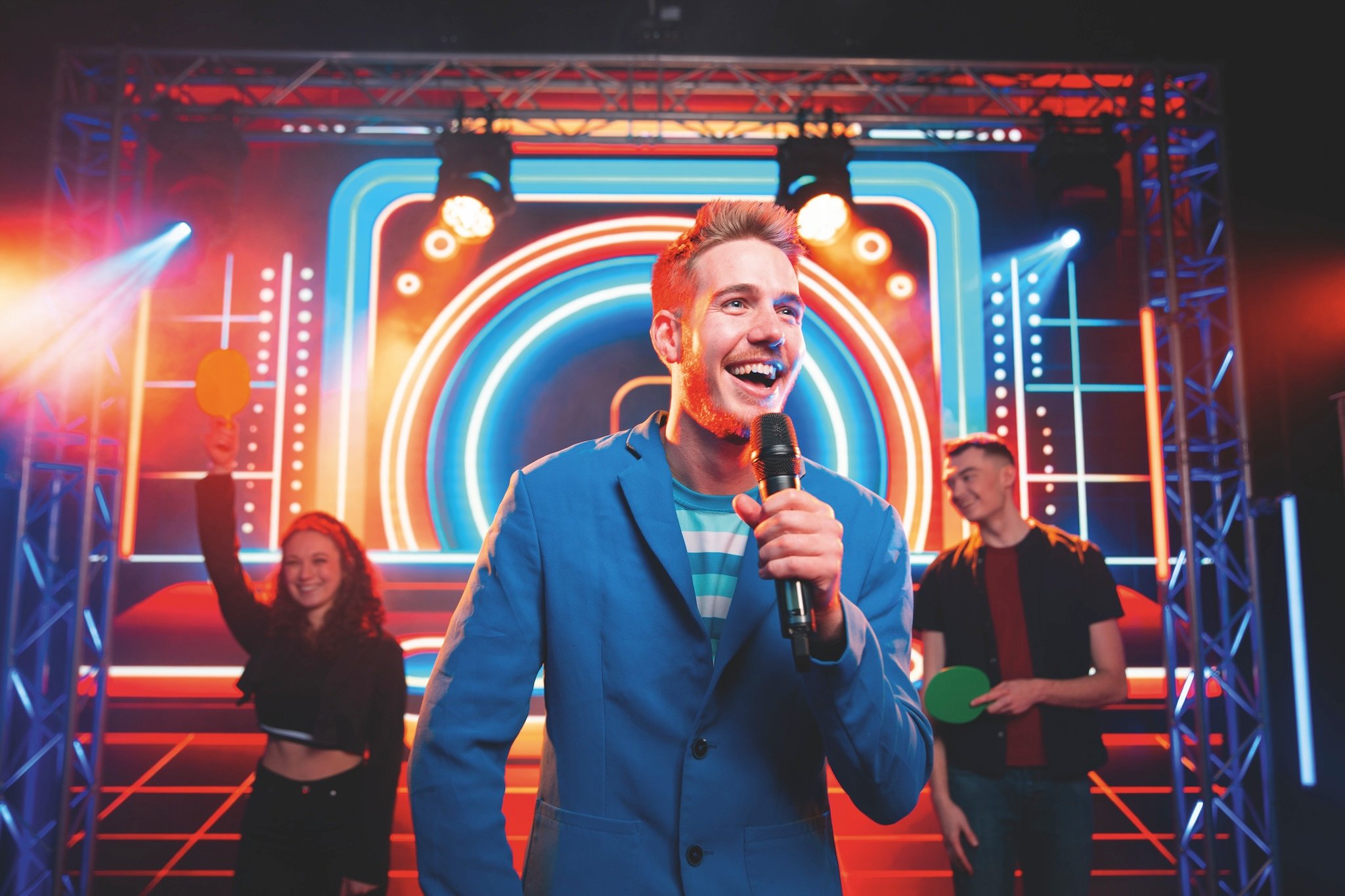A smiling host in a blue blazer holds a microphone on a stage with neon lights and contestants.