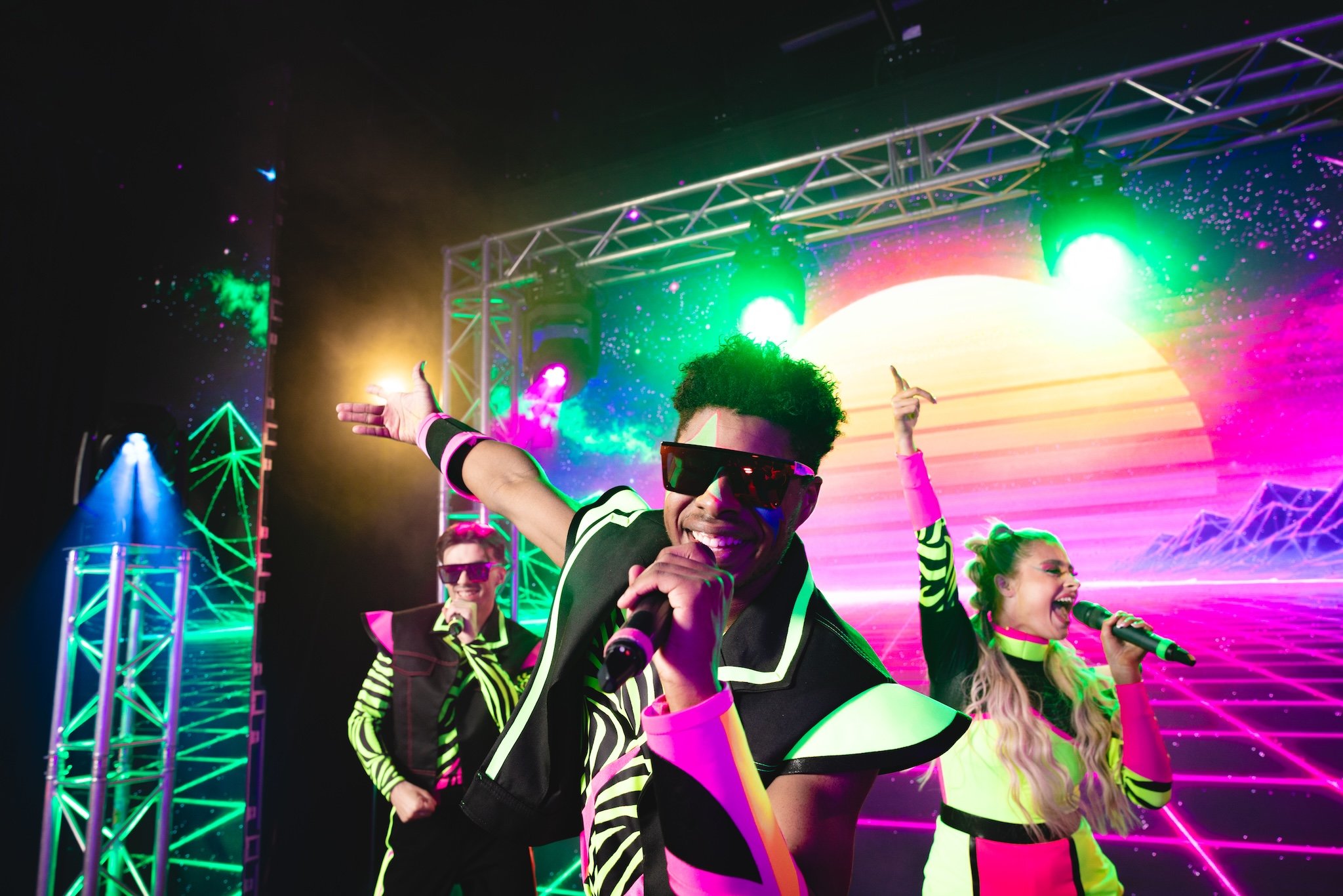 Three vibrant vocalists in retro 80s outfits perform on stage with neon lights and a sunset backdrop.