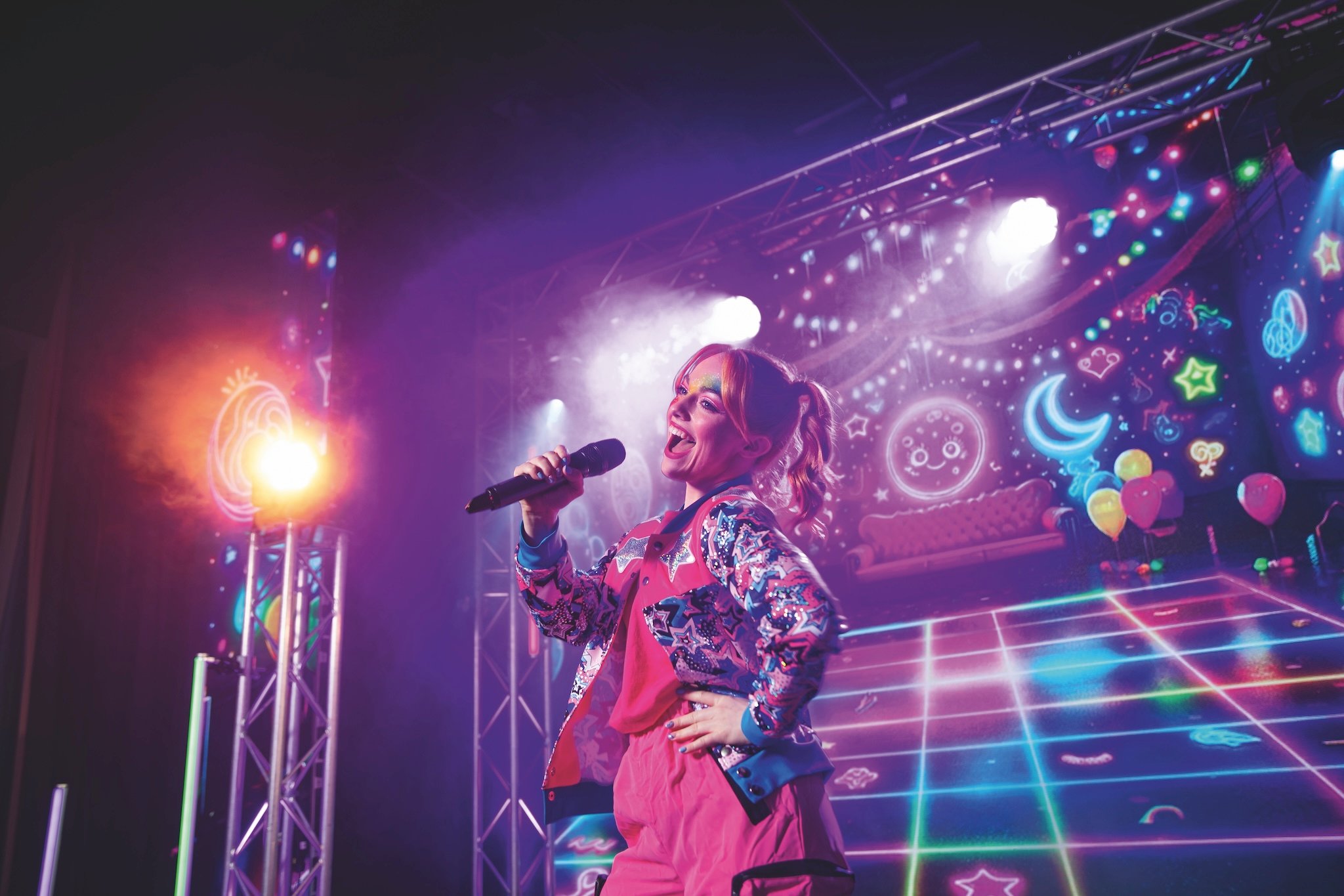 A female artist sings into a microphone on a vibrant stage with glowing neon lights and stage smoke.