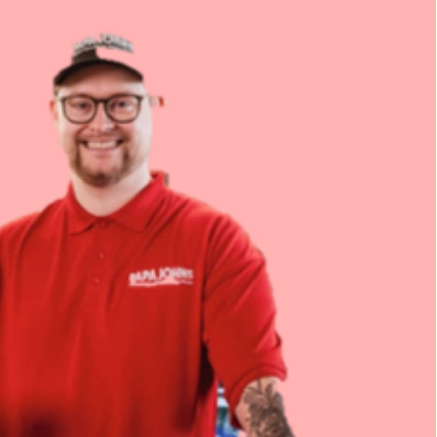 A smiling man wearing a red Papa Johns polo shirt, glasses, and a branded baseball cap against a pink background.