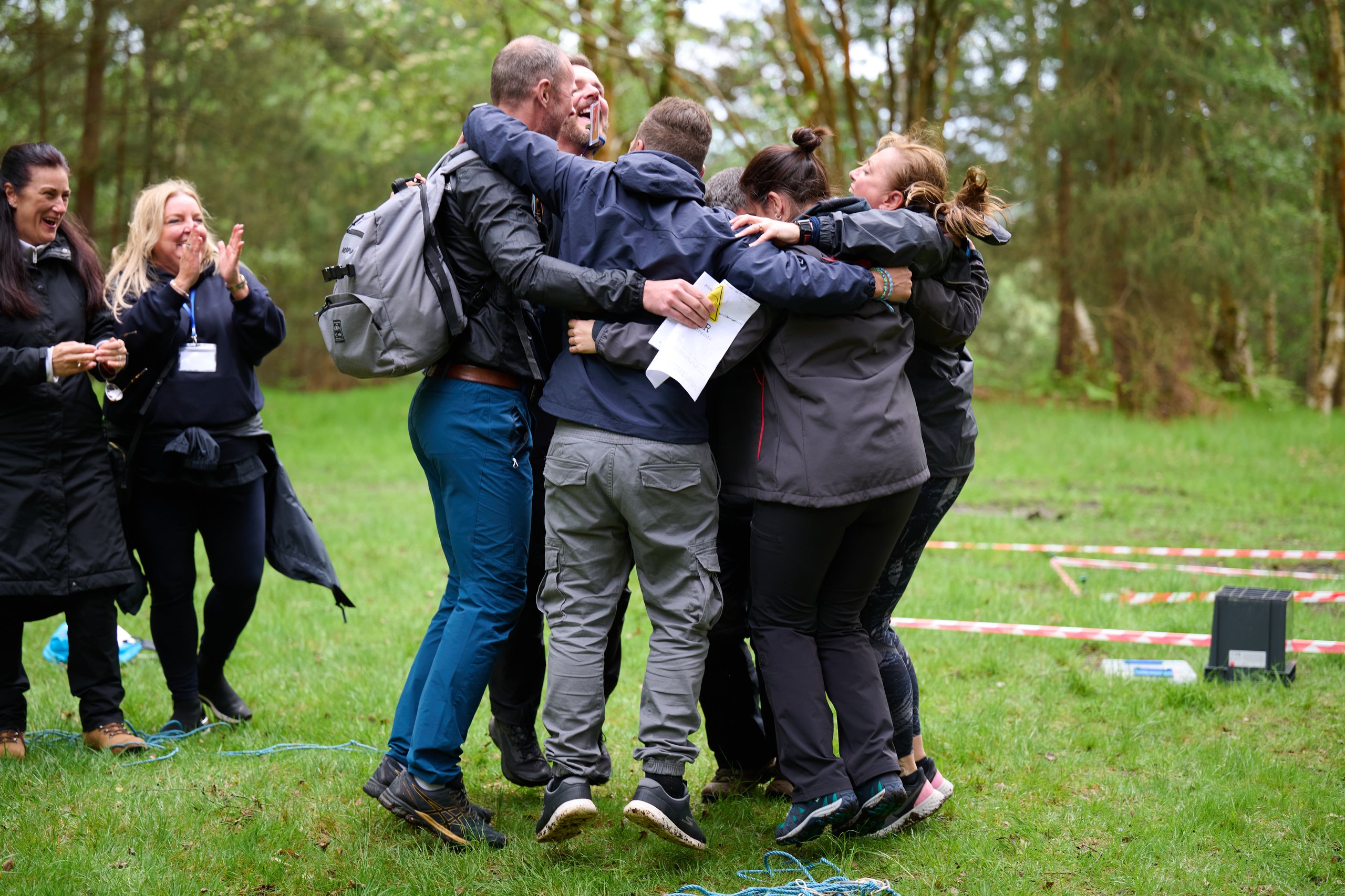 A group of colleagues joyfully hugging and cheering together in a grassy clearing.