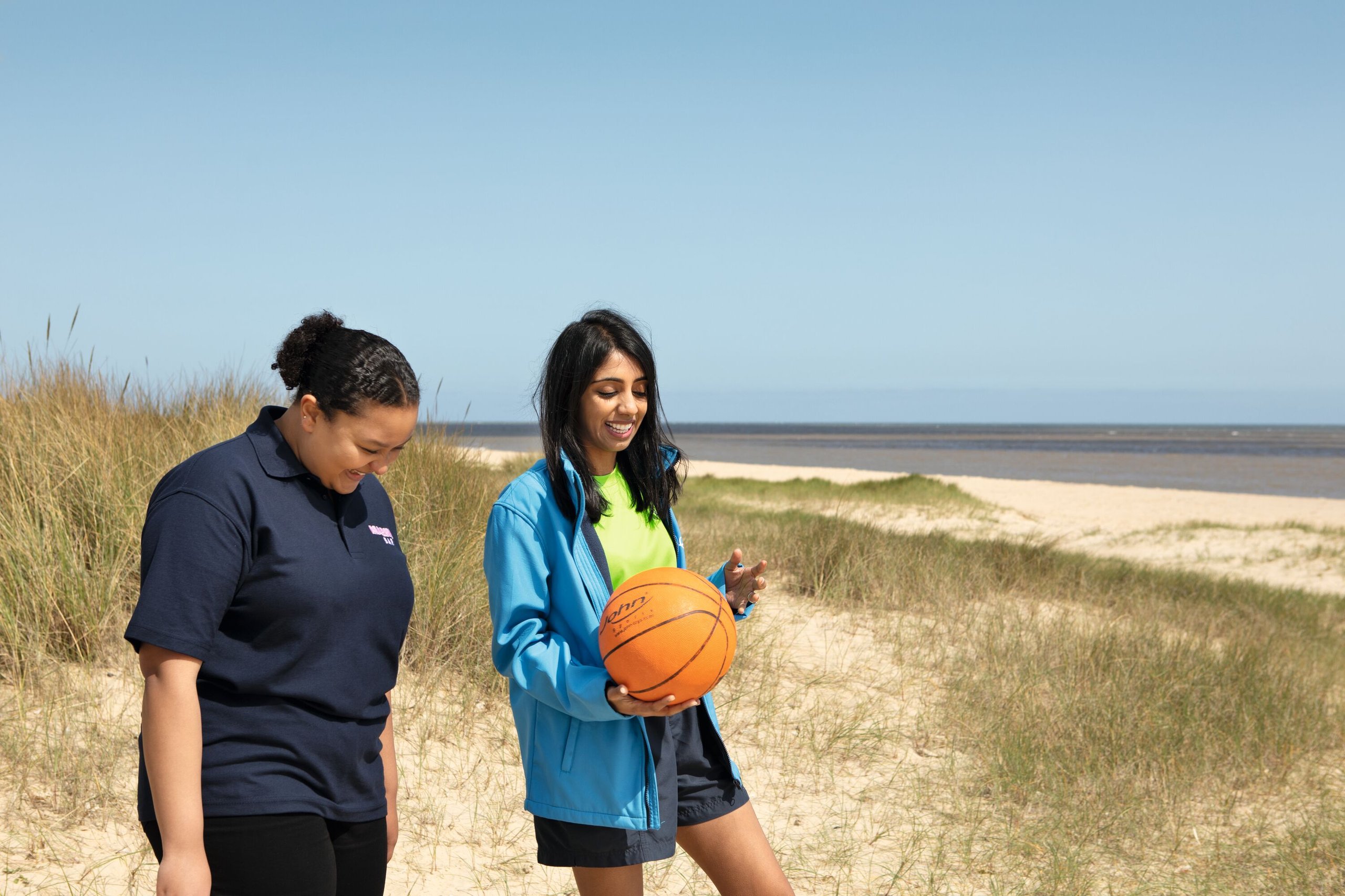 Two young women with a basketball smiling on a beach near grassy dunes under a clear blue sky.