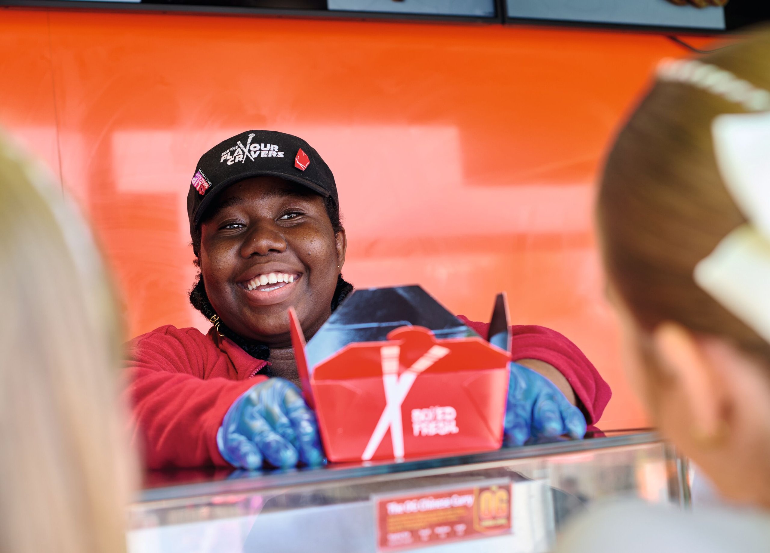 A smiling food vendor in a black hat and red jacket hands a red takeout box to customers.