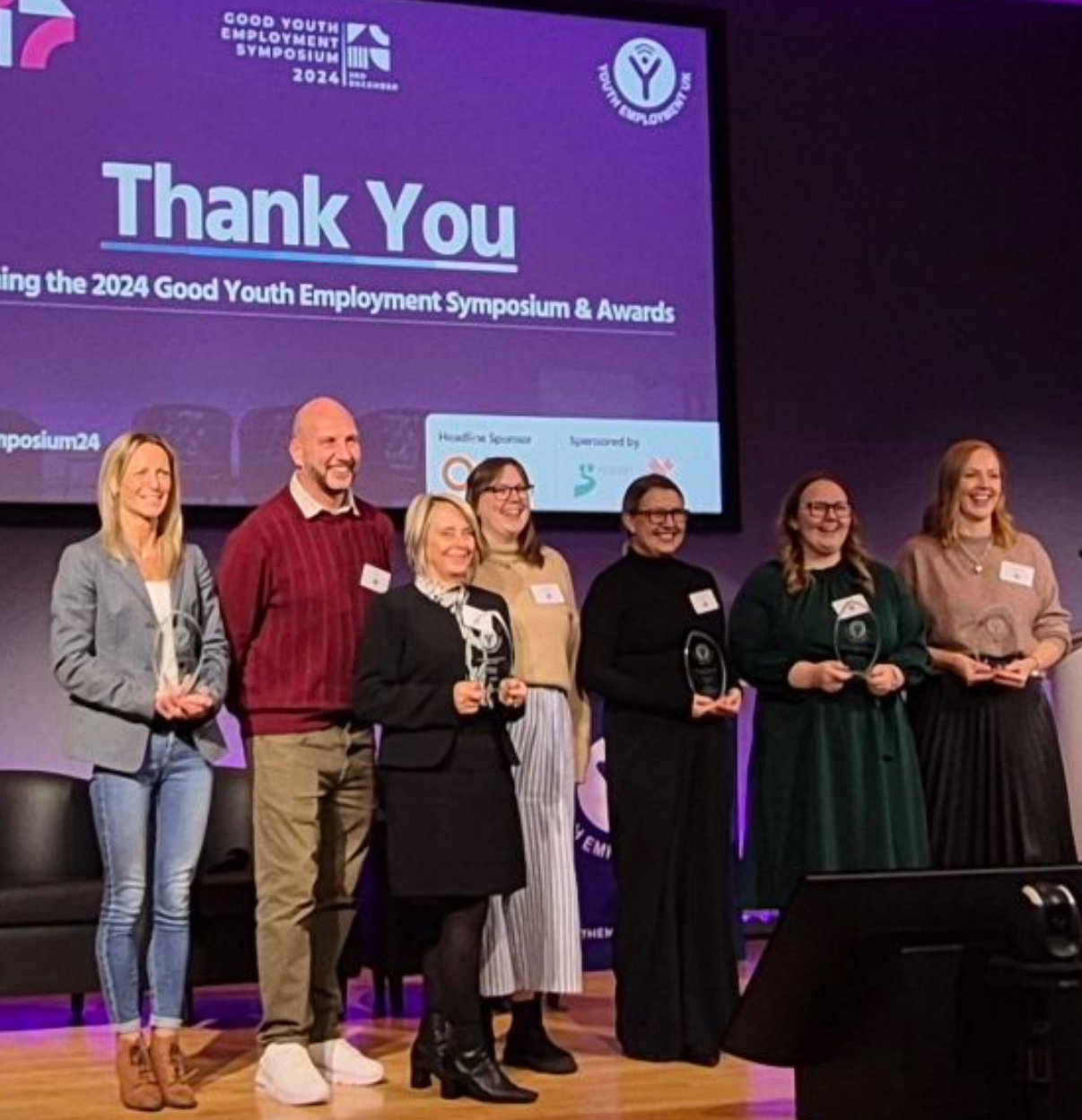 Seven award winners smiling and holding trophies on stage at the 2024 Good Youth Employment Symposium.
